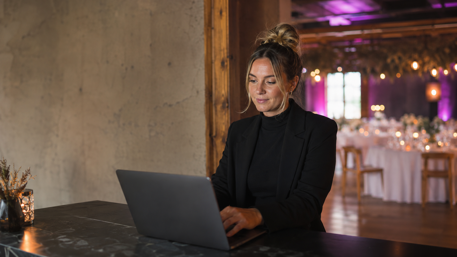 A venue host works at a laptop at a reception desk, a dressed hall with string lights softly blurred behind.