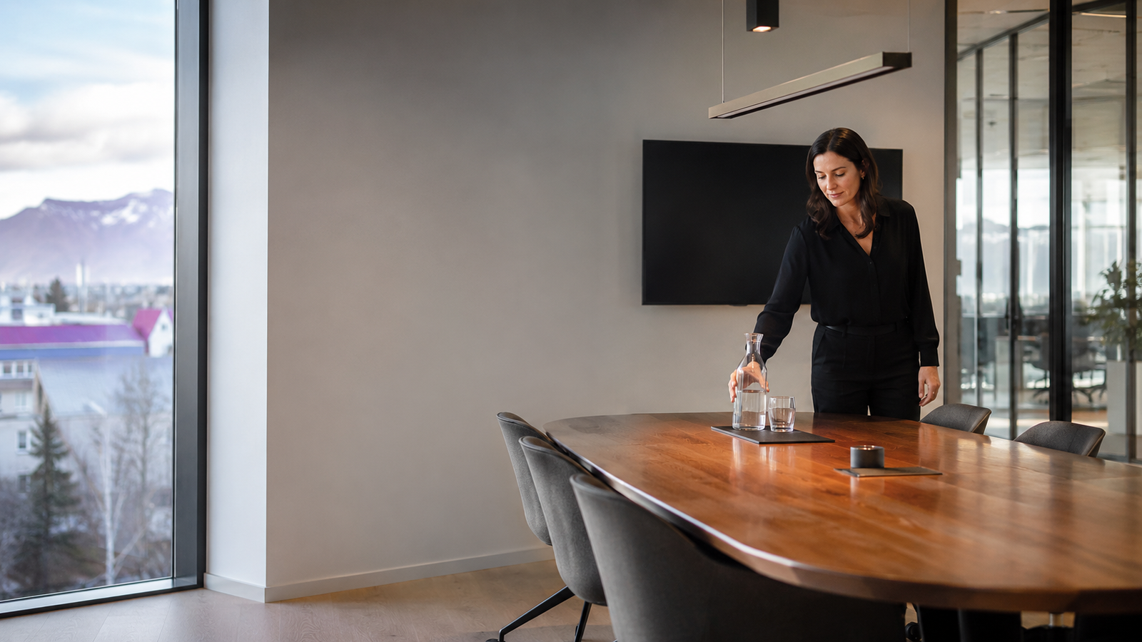 A modern meeting room with a long conference table and a city-and-mountain view; one person sets a water carafe on the table.