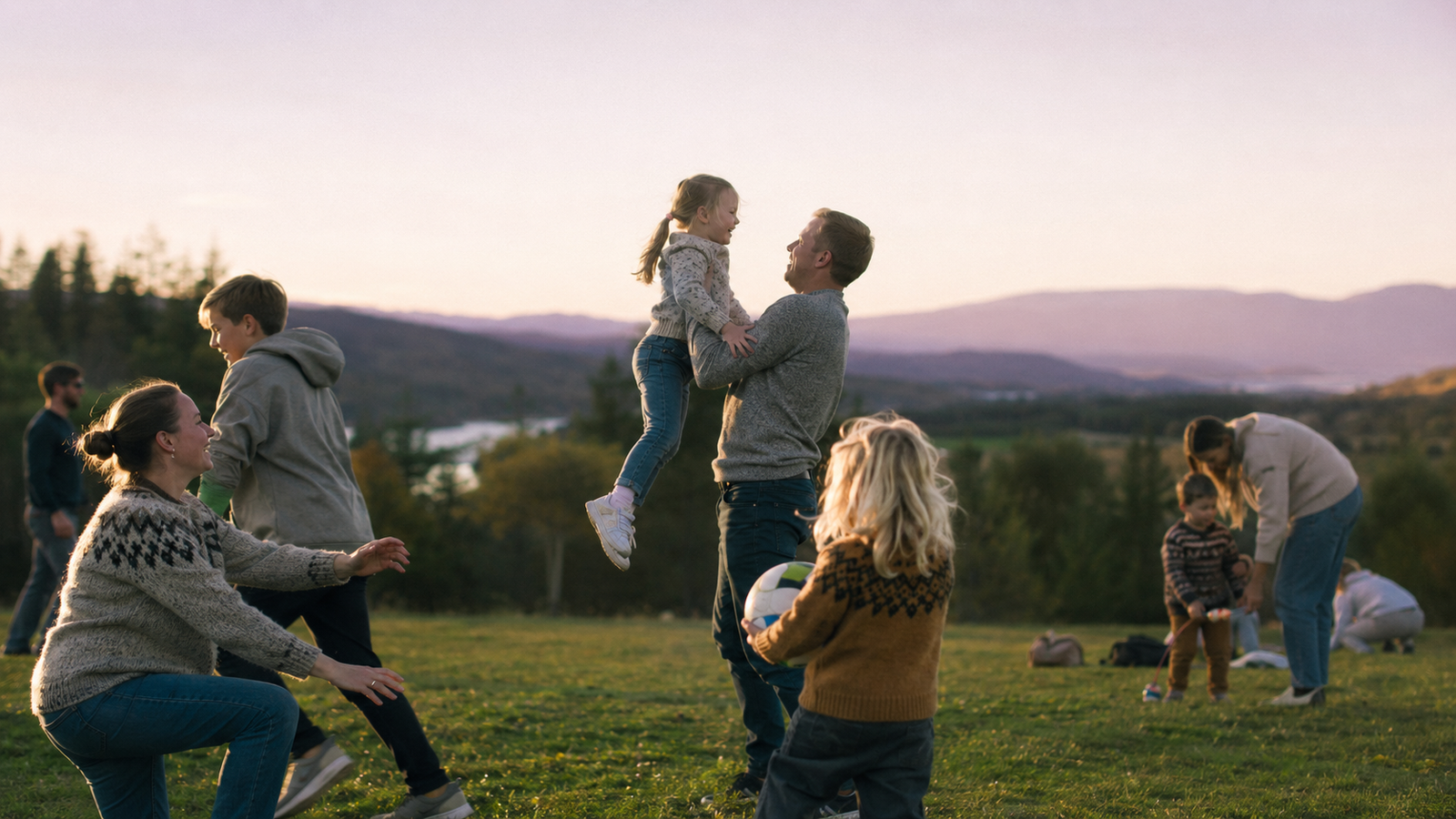 A group of adults and children play together on a grassy field at golden hour with mountains behind them; one adult lifts a child into the air.
