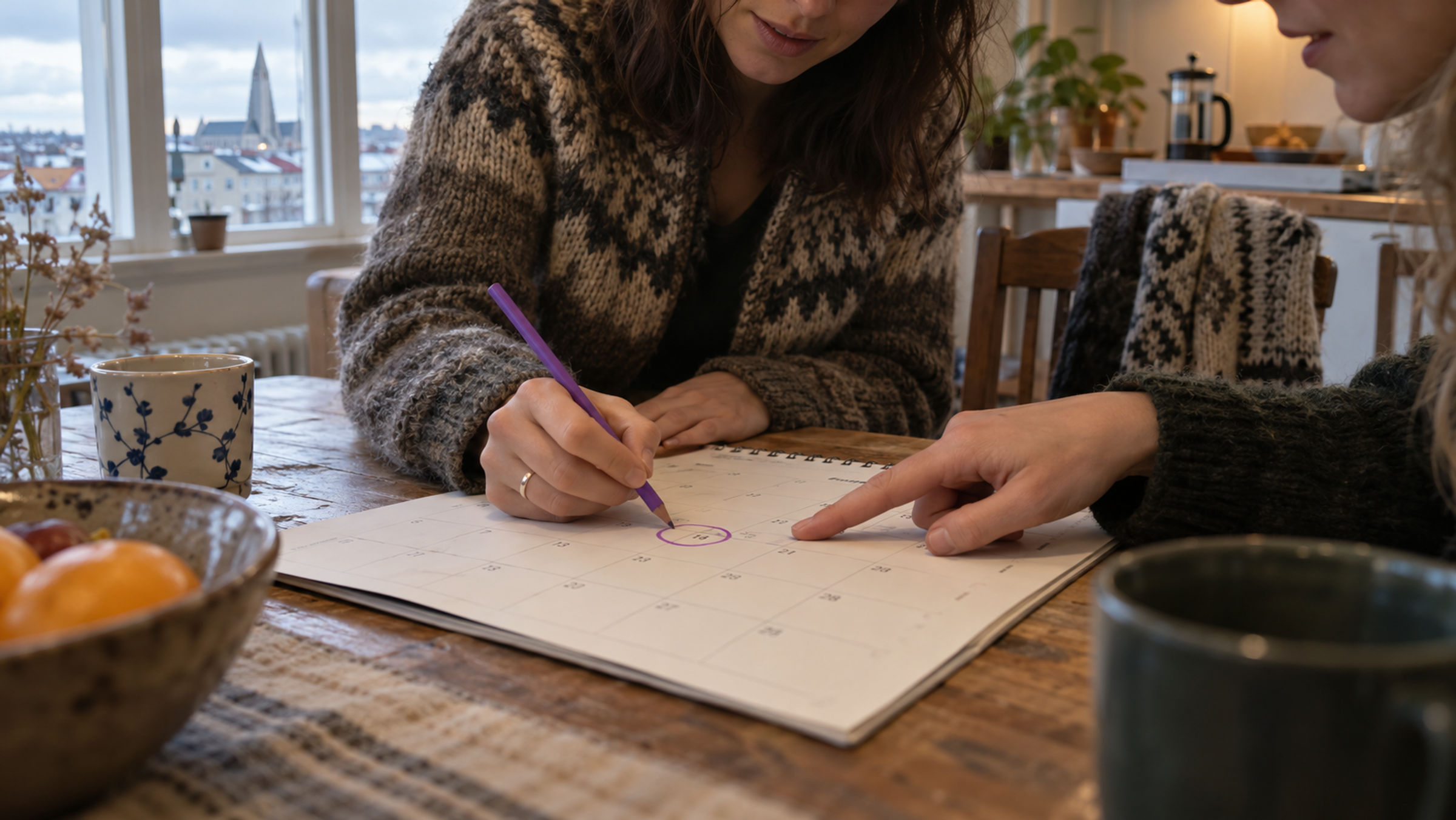 Two friends at a kitchen table in Reykjavík. One circles a single day on a calendar with a purple pencil while the other points to the same day.